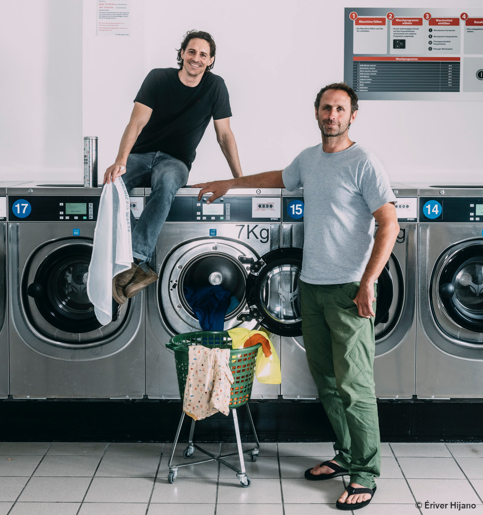 2 Personen in einem Waschraum. Die eine Person sitzt auf den Waschmaschinen mit einem Guppyfriend Waschbeutel in der Hand und die andere Person sitz vor den Waschmaschinen.