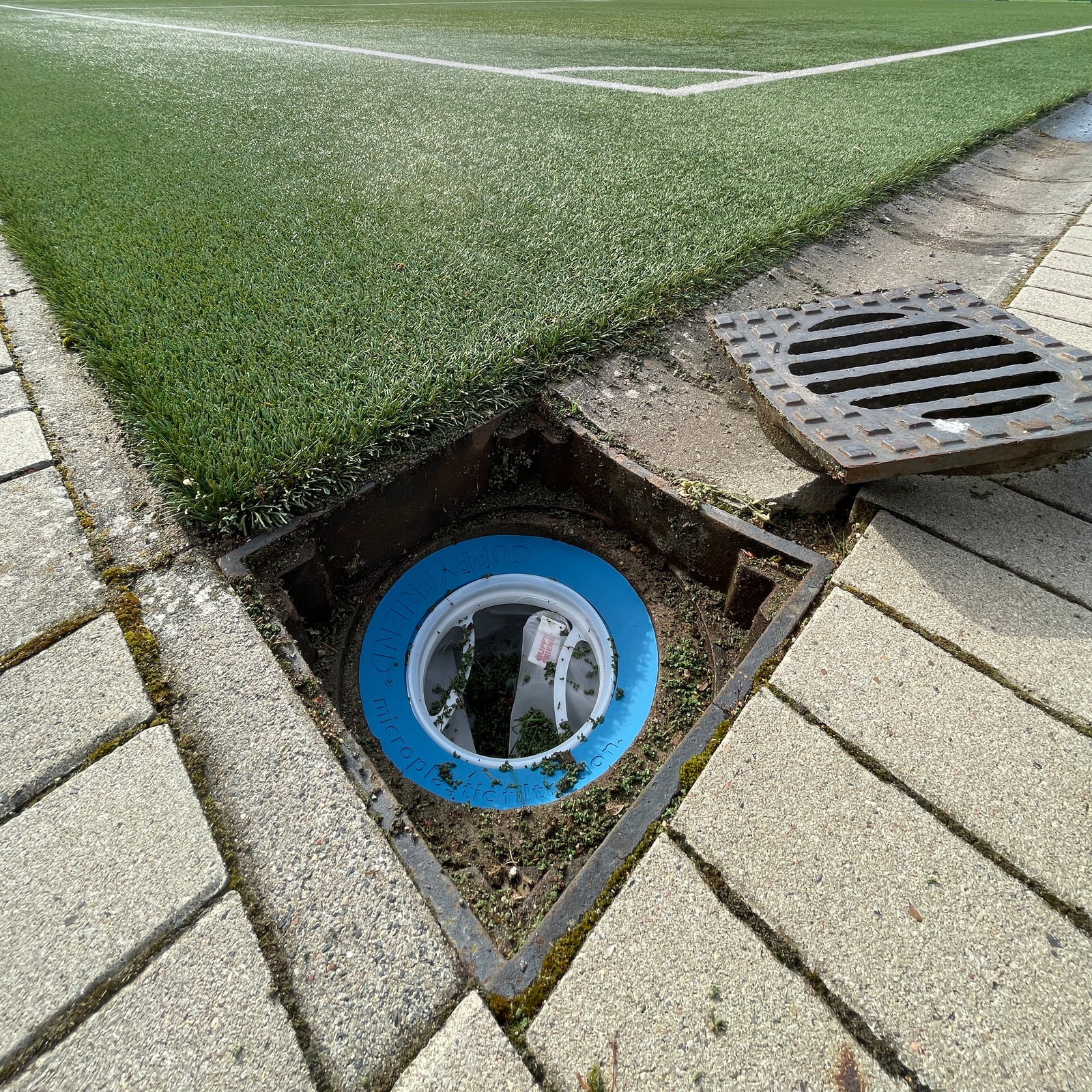 Wasserabfluss an einem Fußballplatz. In dem Abfluss befindet sich ein Guppyfrien Kunstrasen Filter, wo drin sich Infill Material angesammelt hat.
